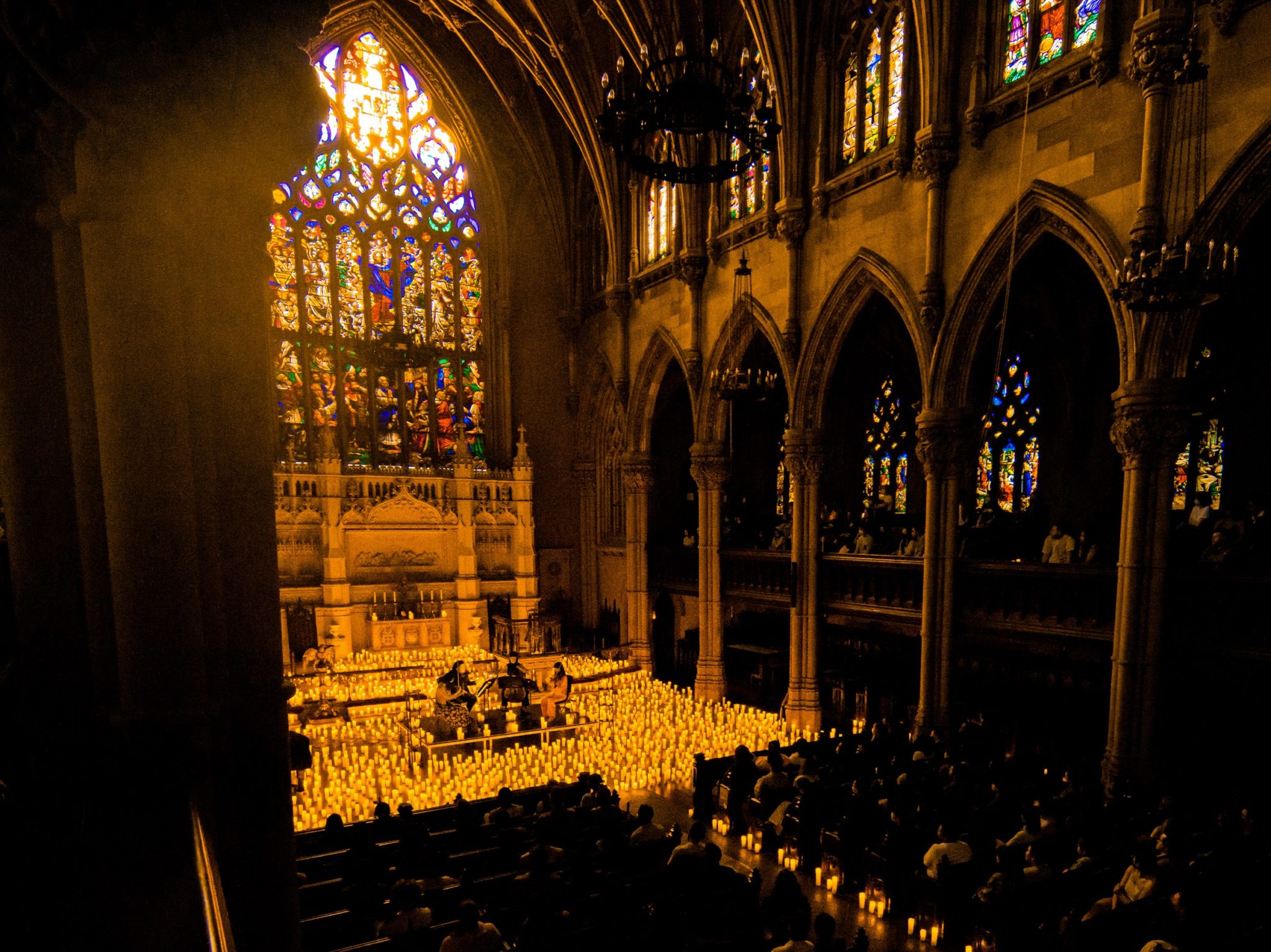 A wide shot of the atlar inside St. Ann's with a string quartet performing surrounded by candles while the pews are filled by the silhouette of an audience.